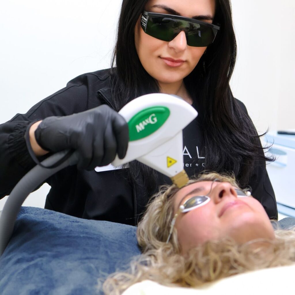 A technician with black hair, protective eyewear and black gloves performs photofacial services on a client's forehead.