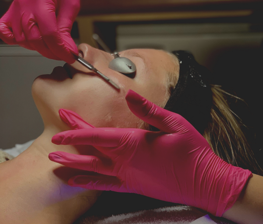 A technician in pink gloves performs dermaplane services on a client's face.