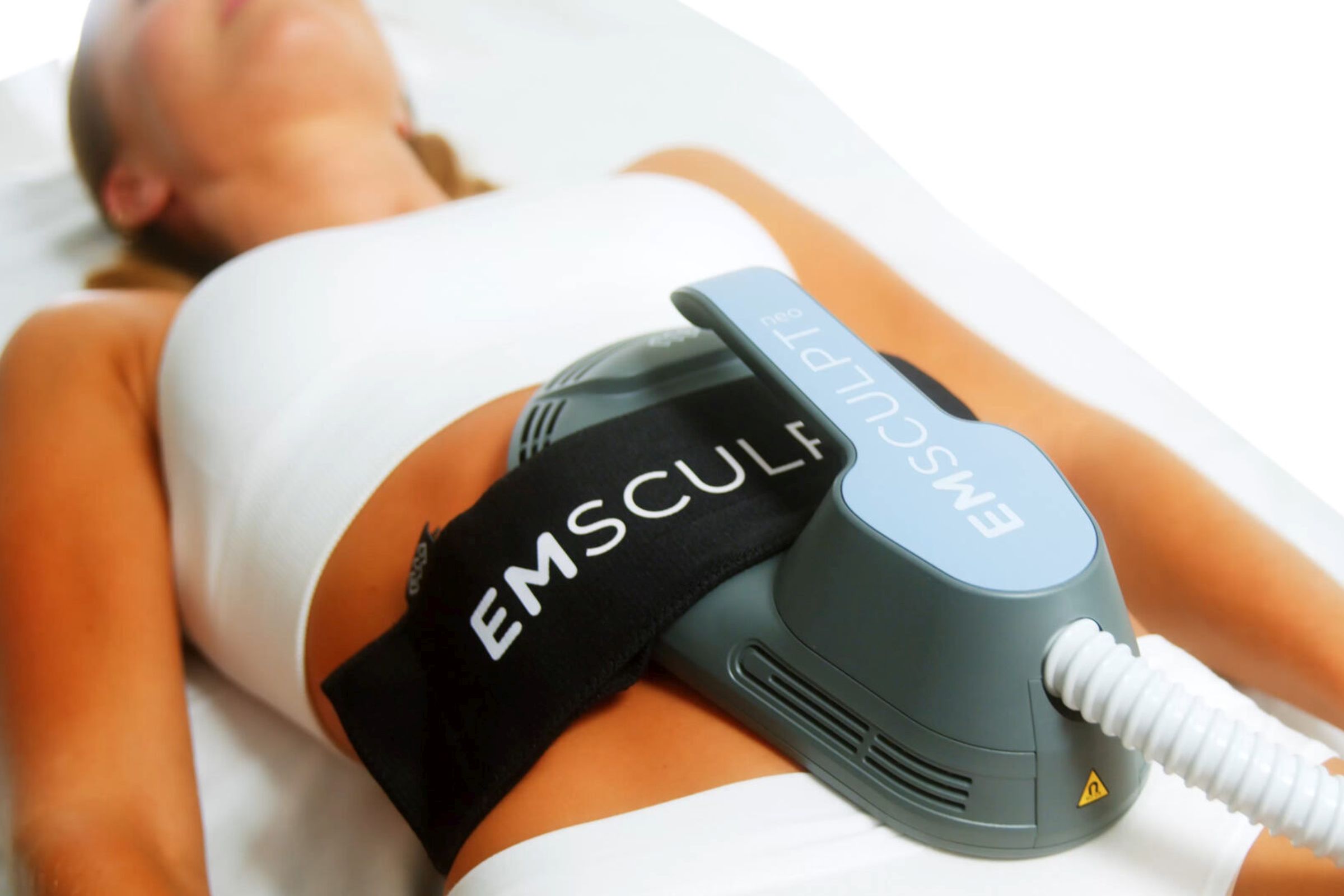 A woman in a white top and bottoms with an Emsculpt NEO device secured to her abdomen.