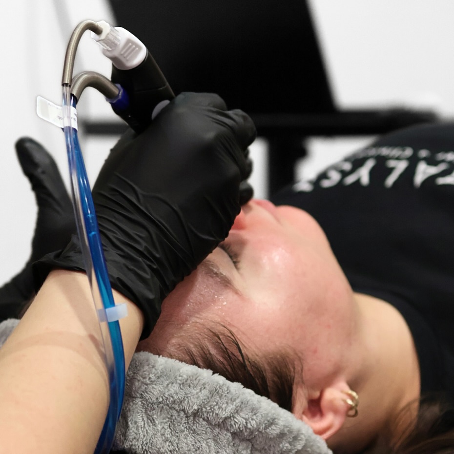 A technician in black gloves performs HydraFacial services on a client's face.